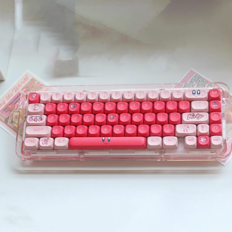 May include: A pink and white mechanical keyboard with custom keycaps. The keyboard has a gradient design, transitioning from white to pink. The keycaps feature character illustrations and the word "Kirby". It is encased in a clear plastic case.