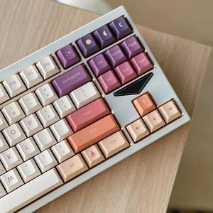 May include: A white mechanical keyboard with a gold trim and pastel pink, purple, and orange keycaps. The keyboard has a full layout with function keys, arrow keys, and a number pad. The keycaps have various symbols and letters printed on them.
