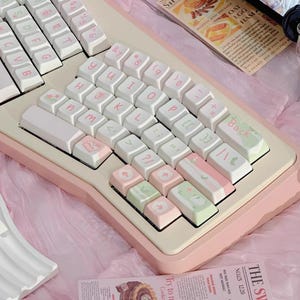 May include: A split ergonomic keyboard with white and pastel pink keycaps. The keyboard has a cream-colored base with pink accents. Some keycaps have decorative icons and text. The keyboard is on a pink surface.