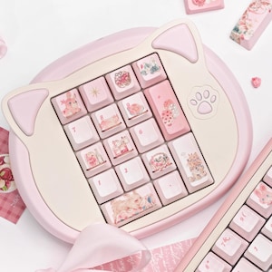 May include: A pink and white cat-shaped keyboard with a paw print keycap. The keyboard has a number pad and a row of keys with various images, including a cat, a star, a flower, and a basket. The keyboard is on a pink and white surface.