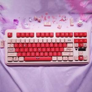 May include: A pink and red mechanical keyboard with custom keycaps. The keyboard has a white base with red and pink keys, some with character illustrations. The word "Kirby" is printed on the space bar.