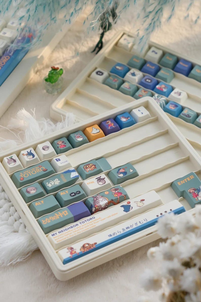 May include: A white plastic keyboard keycap tray with a variety of green, blue, and yellow keycaps featuring illustrations of characters from the movie My Neighbor Totoro. The keycaps are arranged in rows and include the text "My Neighbor Totoro" and "となりのトトロ".