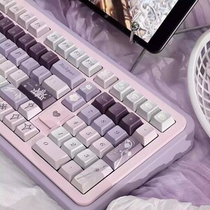 Pode incluir: Teclado mecânico em tons pastel, com base lilás clara e teclas em tons de roxo, lavanda e branco. As teclas apresentam designs e símbolos personalizados, com foco na estética.