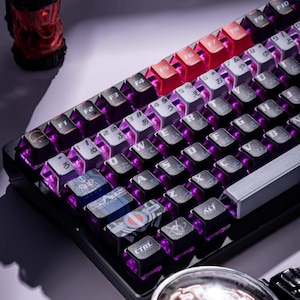 Puede incluir: Un teclado mecánico negro con teclas personalizadas e iluminación púrpura. El teclado presenta teclas rojas y grises, con algunas teclas que muestran diseños gráficos. Las teclas CTRL y ALT son visibles.