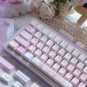 May include: A lilac mechanical keyboard with custom keycaps. The keycaps feature various designs, including hearts, butterflies, and plaid patterns. The keyboard has a light purple frame and white and pink keys. The keyboard is surrounded by flowers and a stuffed animal.