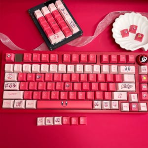 May include: A red mechanical keyboard with pink and white keycaps. The keycaps feature cartoon character designs and the word "Kirby". Additional keycaps are displayed separately. The keyboard is on a red surface.