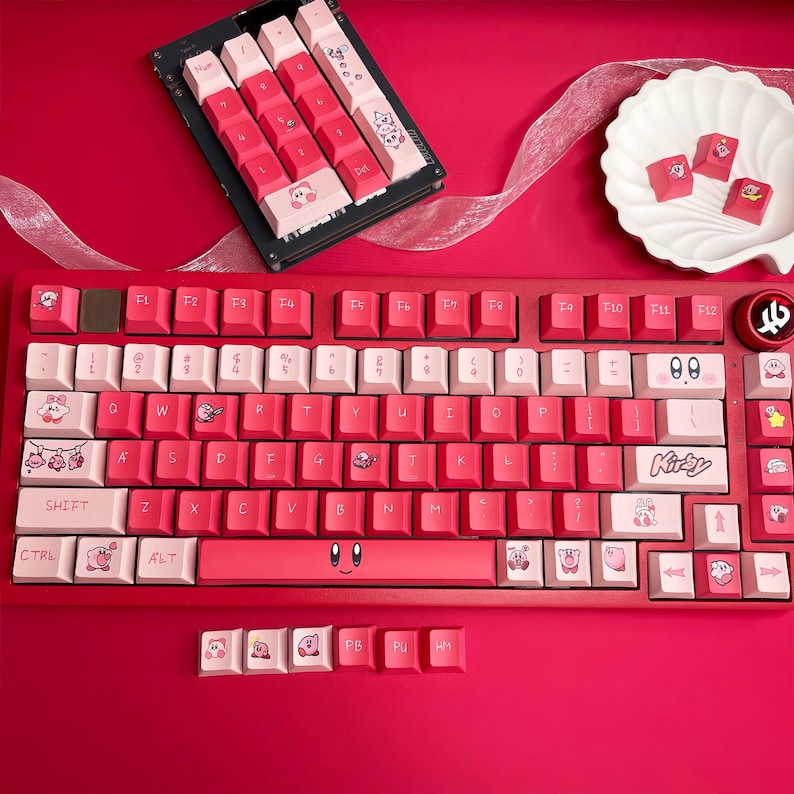 May include: A red mechanical keyboard with pink and white keycaps. The keycaps feature cartoon character designs and the word "Kirby". Additional keycaps are displayed separately. The keyboard is on a red surface.
