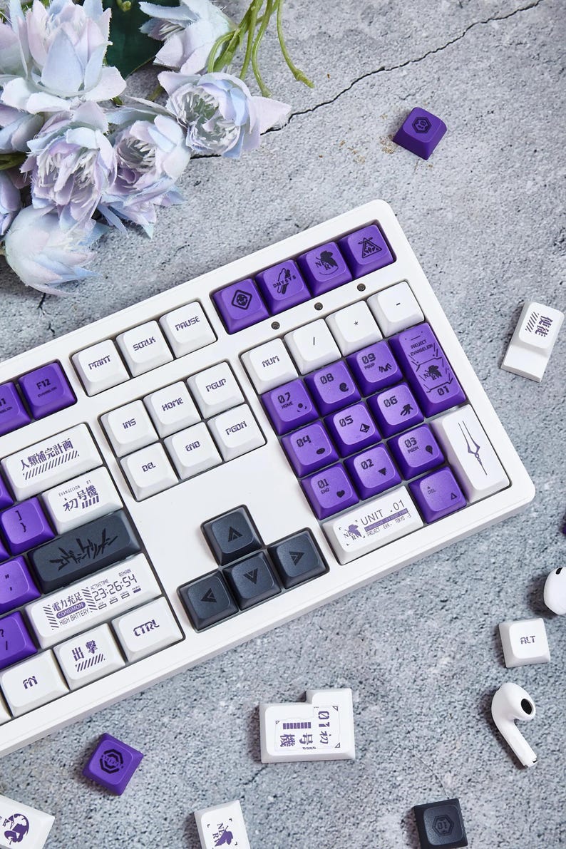 May include: A white mechanical keyboard with purple keycaps featuring Japanese text and symbols. The keyboard has a white and black color scheme and includes arrow keys and function keys. The keyboard is on a gray surface with several purple keycaps scattered around it.