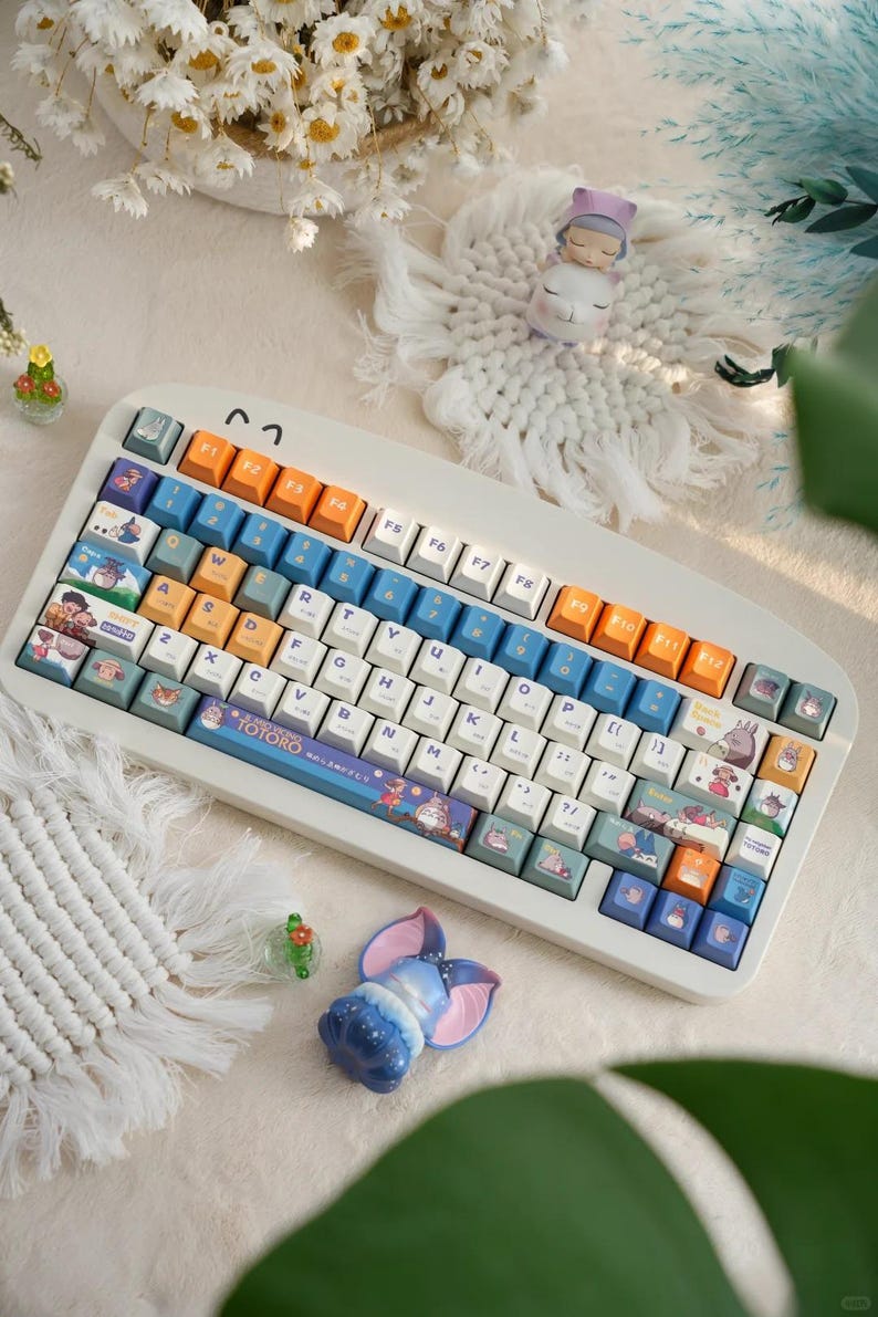 May include: A white mechanical keyboard with custom keycaps featuring characters from the animated film My Neighbor Totoro. The keycaps are in shades of blue, green, and orange. The keyboard has a white and blue spacebar with the text "My Neighbor Totoro" printed on it.