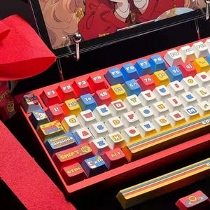 May include: A red and yellow keyboard with colourful keycaps featuring cartoon characters and symbols. The keyboard has a red frame and a yellow and blue stripe design. The keys are white with black lettering. The keyboard has a row of function keys labelled F1 through F11.