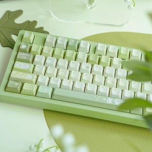 Puede incluir: Un teclado mecánico verde con teclas blancas y un marco verde claro. Las teclas tienen un diseño floral y el teclado tiene una barra espaciadora con un diseño floral.