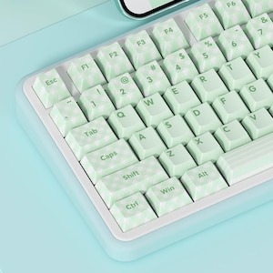 Può includere: Una tastiera bianca con tasti verdi e un motivo a scacchi. I tasti hanno lettere e simboli stampati su di essi. La tastiera è su una superficie blu chiaro.