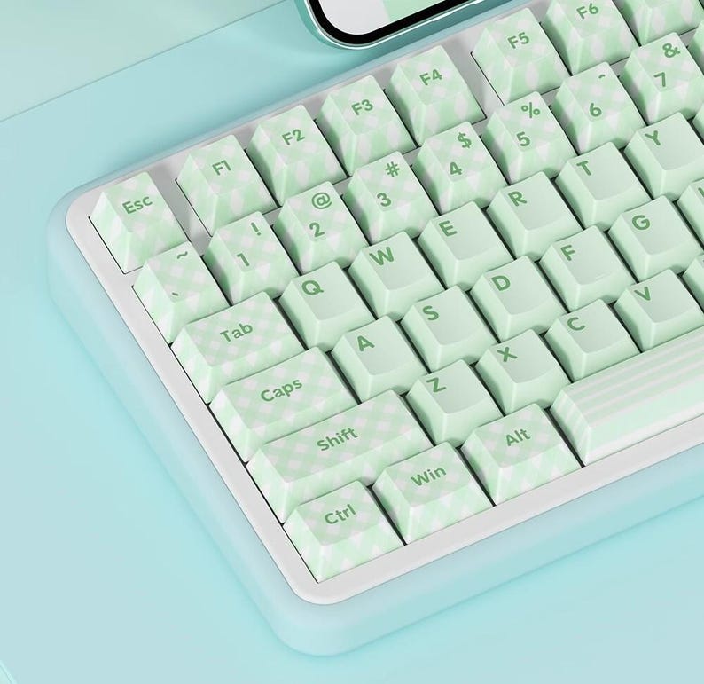 May include: A white keyboard with green keys and a checkered pattern. The keys have letters and symbols printed on them. The keyboard is on a light blue surface.