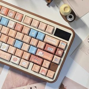 May include: A brown and white keyboard with a variety of keycaps featuring cute illustrations of animals, clouds, and other designs. The keyboard has a Japanese layout with keys labeled in Japanese characters.
