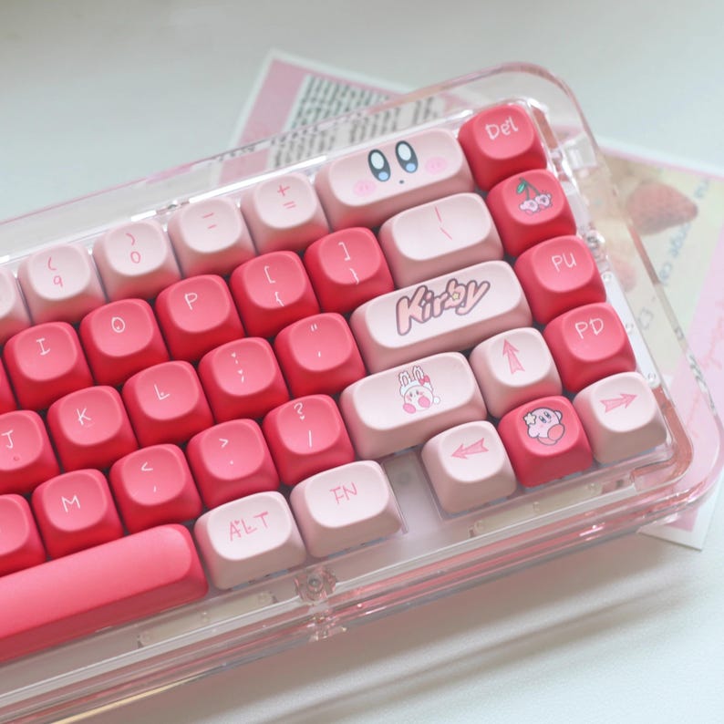 May include: A pink and red mechanical keyboard with custom keycaps. The keycaps feature the word "Kirby" and various cartoon character designs. The keyboard is housed in a clear plastic case.