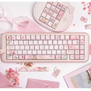 May include: A pink mechanical keyboard with floral and pastel-colored keycaps. The keyboard has a pink frame and a white keycap base. The keyboard is surrounded by pink and white decorative items.