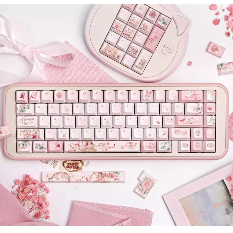 May include: A pink mechanical keyboard with floral and pastel-colored keycaps. The keyboard has a pink frame and a white keycap base. The keyboard is surrounded by pink and white decorative items.