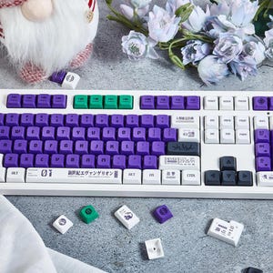 May include: A white mechanical keyboard with purple and green keycaps. The keyboard has a Japanese language layout and the text "01 暁光とエヴァンゲリオン" is printed on the bottom row of keys.