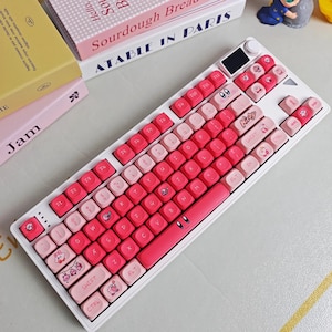 May include: A white mechanical keyboard with pink and light pink keycaps. The keycaps feature cartoon character designs and the word "Kirby". The keyboard has a small screen and a rotary dial. The keyboard is surrounded by books.