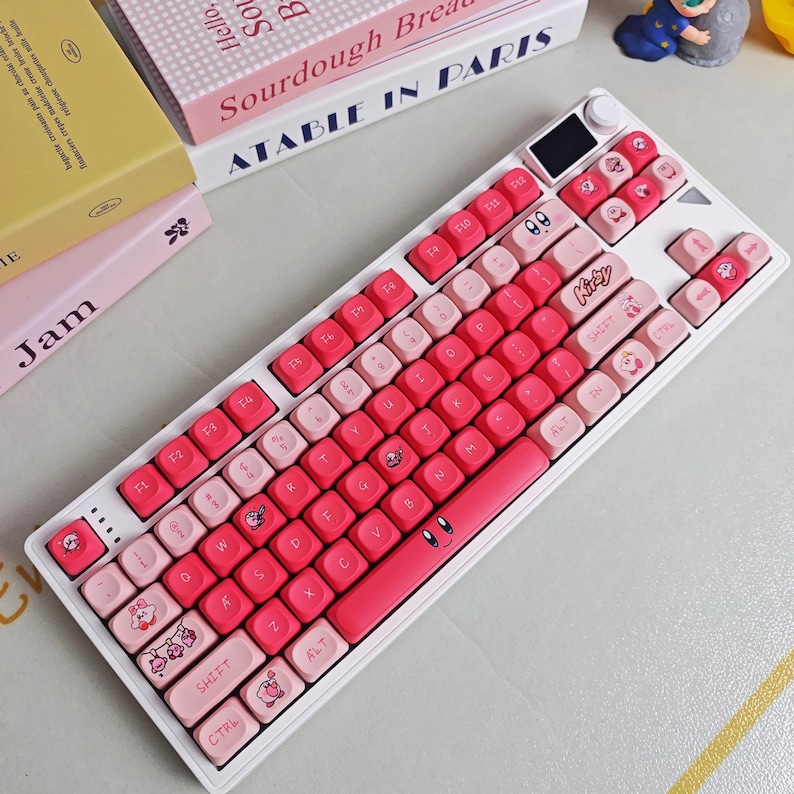 May include: A white mechanical keyboard with pink and light pink keycaps. The keycaps feature cartoon character designs and the word "Kirby". The keyboard has a small screen and a rotary dial. The keyboard is surrounded by books.