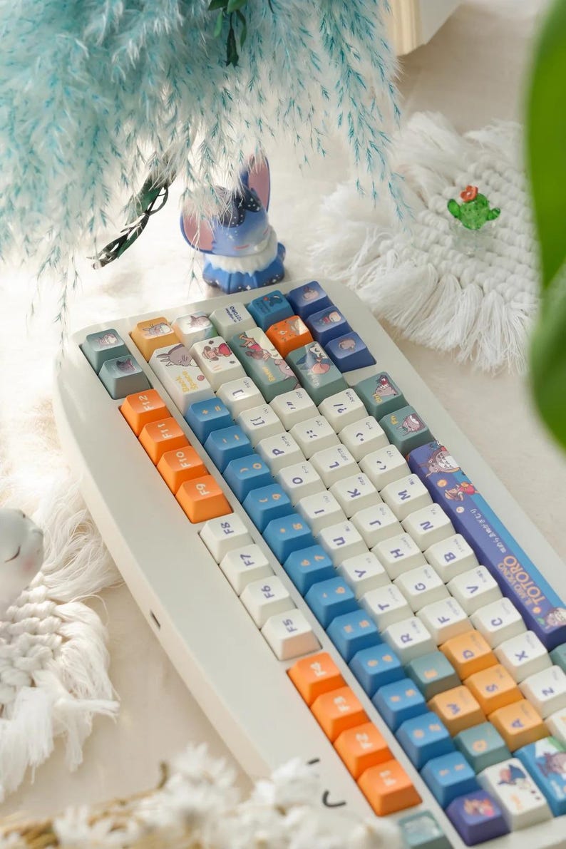 May include: A white keyboard with blue, orange, and white keycaps. The keycaps have illustrations of cartoon characters. The keyboard has a white cable and a white USB connector. The keyboard is on a white surface.