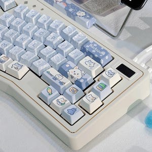 Puede incluir: Un teclado mecánico blanco y dorado con teclas azul claro. Las teclas presentan ilustraciones de gatos e iconos de comida. El teclado tiene un reposamuñecas a juego con un diseño similar. El teclado está sobre una superficie blanca.