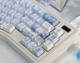 May include: A white and gold mechanical keyboard with light blue keycaps. The keycaps feature cat illustrations and food icons. The keyboard has a matching wrist rest with a similar design. The keyboard is on a white surface.