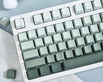 May include: A white mechanical keyboard with green keycaps. The keyboard has a standard layout with function keys, number keys, and a space bar. The keycaps are a gradient of green, with the darker green keys being on the bottom row and the lighter green keys being on the top row.