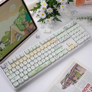 May include: A white keyboard with pastel-colored keys and cartoon animal keycaps. The keyboard has a green and white keycap set with a cartoon camping scene. The keyboard is on a white surface with a tablet and flowers in the background.