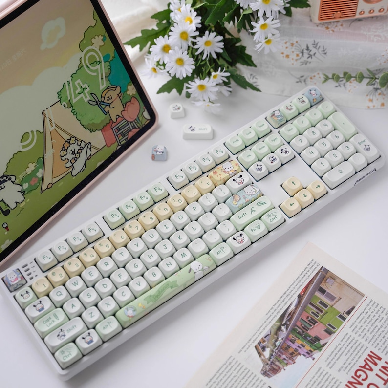 May include: A white keyboard with pastel-colored keys and cartoon animal keycaps. The keyboard has a green and white keycap set with a cartoon camping scene. The keyboard is on a white surface with a tablet and flowers in the background.