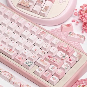 May include: A pink mechanical keyboard with custom keycaps featuring a variety of pastel-colored designs, including strawberries, flowers, and ribbons. The keycaps are labeled with letters and numbers, and the keyboard has a pink frame.