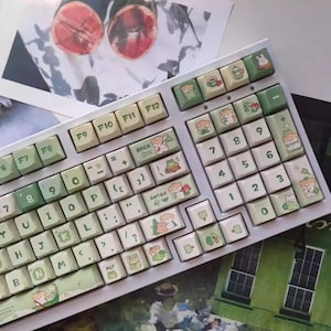 May include: A white mechanical keyboard with custom keycaps. The keycaps feature a light green and cream color scheme with illustrations of bears, mushrooms, and other nature-themed designs. The keyboard has a standard layout with function keys.