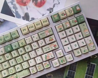 May include: A white mechanical keyboard with custom keycaps. The keycaps feature a light green and cream color scheme with illustrations of bears, mushrooms, and other nature-themed designs. The keyboard has a standard layout with function keys.
