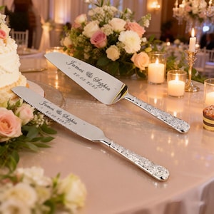 May include: Silver cake server and knife set with engraved names "James & Sophia" and date "20-05-2025". The handles have a decorative floral pattern. The set is displayed on a table with flowers and candles.