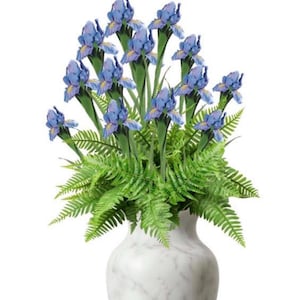 May include: A floral arrangement featuring blue iris flowers and green fern foliage in a white marble-patterned vase. The irises have a delicate, layered petal design. The vase has a classic, elegant shape.