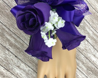 May include: Purple silk rose wrist corsage with a purple satin bow and white baby's breath.