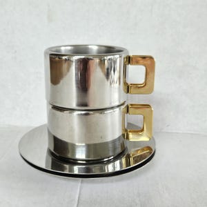 May include: A set of two stacked cylindrical silver-toned metal cups resting on a matching circular saucer. Each cup features a distinct, square-shaped gold-toned handle attached to the side, creating a contrasting metallic aesthetic.