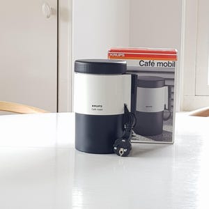 Puede incluir: Una cafetera Krups Café mobil en blanco y negro con su caja. La cafetera tiene un diseño cilíndrico con la parte superior e inferior negras y el centro blanco. La caja presenta el logotipo de Krups y las palabras "Café mobil".