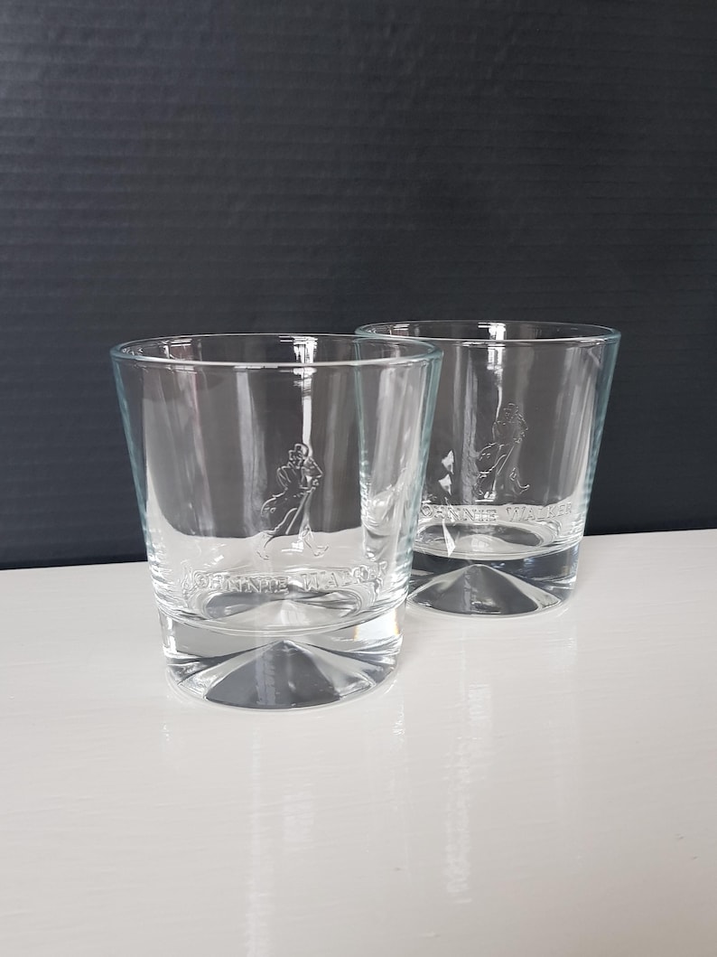 K&ouml;nnte beinhalten: Zwei klare, kurze Whiskygl&auml;ser. Jedes Glas hat ein Logo eines Mannes mit Gehstock und die Worte "JOHNNIE WALKER" auf der Vorderseite eingraviert. Die Gl&auml;ser stehen auf einer wei&szlig;en Oberfl&auml;che mit dunklem Hintergrund.