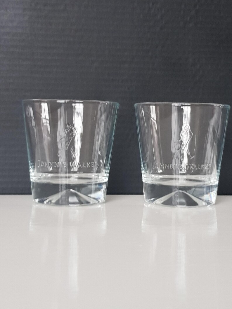 K&ouml;nnte beinhalten: Zwei klare Whiskeygl&auml;ser mit dem eingravierten "Johnnie Walker"-Logo auf der Vorderseite. Die Gl&auml;ser sind zylindrisch mit leicht ausgestellter Oberseite und stehen auf einer wei&szlig;en Oberfl&auml;che vor einem dunklen Hintergrund.