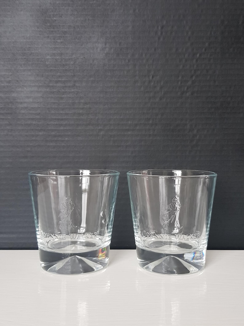 K&ouml;nnte beinhalten: Zwei transparente Whiskeygl&auml;ser mit dem Johnnie Walker-Logo auf der Vorderseite. Die Gl&auml;ser haben einen dicken Boden und ein gerades Design. Der Hintergrund ist eine dunkelgraue, strukturierte Oberfl&auml;che.
