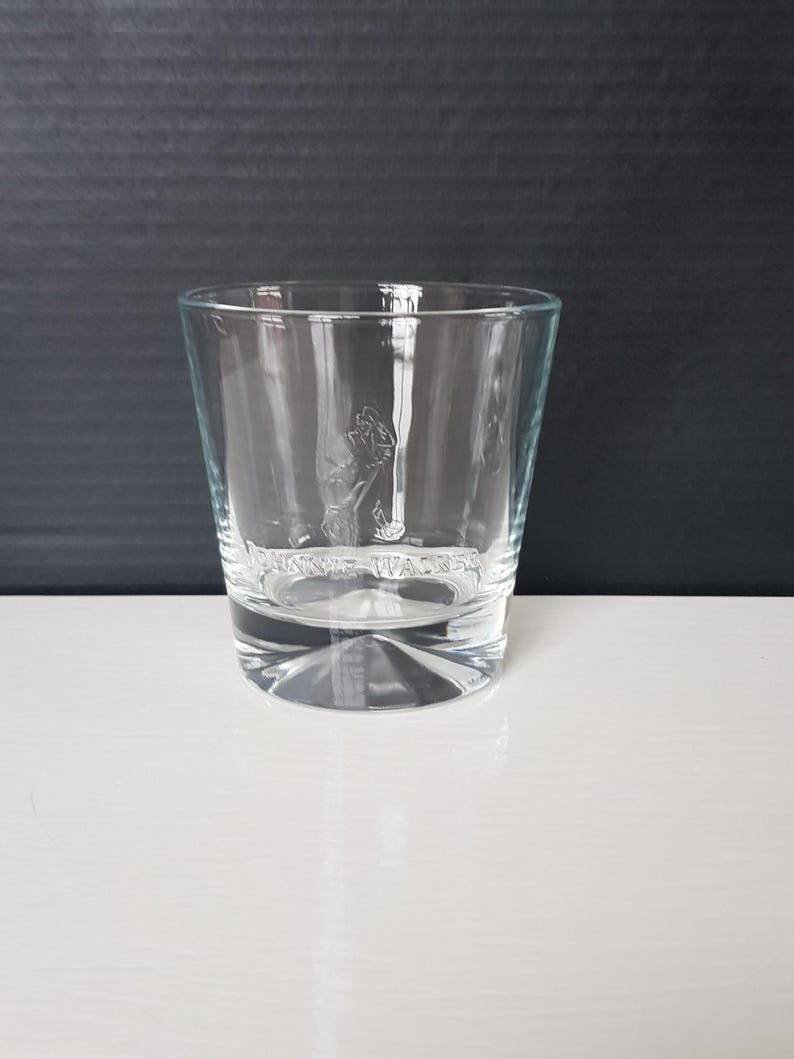 K&ouml;nnte beinhalten: Ein klares, kurzes Trinkglas mit dickem Boden. Das Glas zeigt ein ge&auml;tztes Bild eines Mannes, der geht, und die Worte "JOHNNIE WALKER" darunter. Das Glas steht auf einer wei&szlig;en Oberfl&auml;che und einem dunklen Hintergrund.