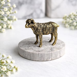 May include: A detailed, golden-colored ram figurine stands on a small, round, gray marble base. The ram has textured fur and detailed horns. The background is blurred, with white flowers and a marble-like surface.