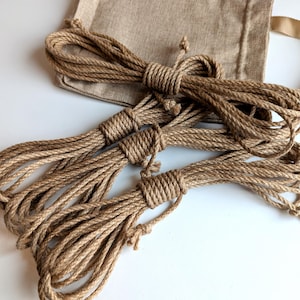 Puede incluir: Tres bobinas de cuerda de yute trenzada, gruesa y marrón, dispuestas en una fila horizontal, cada una asegurada con un nudo central. Una bolsa de lino de color tostado claro, rectangular y con cordón, con un asa a juego, se sitúa detrás.