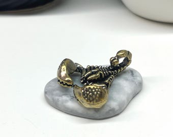May include: A gold-colored scorpion figurine with intricate details. The scorpion is posed with its tail raised and its claws extended. The figurine is sitting on a white marble-like surface.