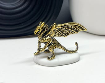 May include: A small gold dragon figurine with black detailing, perched on a white marble base. The dragon has wings spread out and a long tail.