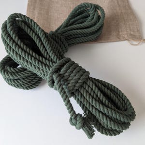 Puede incluir: Una longitud de cuerda de color verde oliva oscuro enrollada con una textura trenzada gruesa. La cuerda está dispuesta en múltiples bucles y presenta un extremo anudado con una pequeña borla deshilachada. Se ve una bolsa de tela beige.