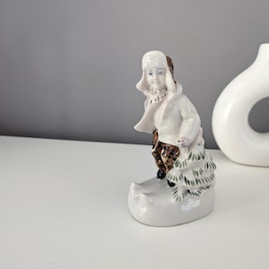 May include: A porcelain figurine of a child in winter attire. The figure is wearing a white coat, hat, and scarf, with brown patterned pants. The figurine is standing on a snow-covered base with a small fir tree.
