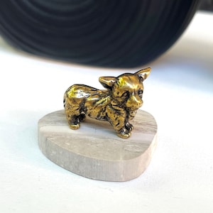 May include: A small, golden-colored dog figurine. The dog is standing on a heart-shaped, light gray stone base. The figurine has detailed features and a textured surface. The background includes a black and white object.