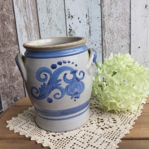 May include: A vintage ceramic pot with a blue and white floral design. The pot has two handles and a blue rim. The pot is sitting on a cream-colored lace doily.